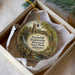 A decorative ornament in a white box, featuring a painting of a historical ironworks facility surrounded by trees and a bridge.