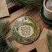 A decorative ornament featuring a scenic image of Tannenhill Ironworks Historical State Park, Alabama, with a waterfall and a bridge. The ornament is placed on a wooden surface, surrounded by pine branches and a cup of coffee.