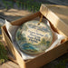 A decorative ornament in a wooden box, featuring an illustration of a scenic landscape with palm trees and a body of water.