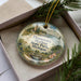 A decorative ornament in a box, featuring a scenic landscape with a lighthouse and a bridge, surrounded by pine branches.
