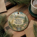 A decorative ornament featuring a lighthouse and a scenic landscape, placed on a wooden surface alongside a cup of coffee and some pine branches.