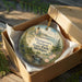 A decorative ornament in a wooden box, featuring a lighthouse and a scenic landscape, with the text "Thomson's Harbor State Park, Michigan" printed on it.