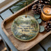 A decorative ornament with a scenic image of Tippecanoe River State Park, Indiana, is displayed on a wooden tray alongside pine cones and a lit candle.