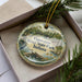 A decorative ornament in a box, featuring a scenic image of Tippecanoe River State Park, Indiana.