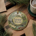 A decorative ornament featuring a scenic image of Tishomingo State Park, Mississippi, with a waterfall and a bridge, is displayed on a wooden surface alongside a cup of coffee and some pine branches.