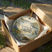 A decorative ornament in a wooden box, featuring a scenic landscape with a lake, mountains, and wildlife.