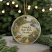 A round ornament with a painting of a park scene, featuring a bird, a boat, and a bridge, hanging in front of a Christmas tree with lights.