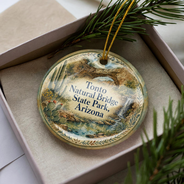 A decorative ornament with a landscape scene featuring a bridge, cactus, and water, placed in a box with pine branches.