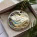 A decorative ornament in a box, featuring a scenic landscape with mountains, trees, and a body of water.