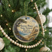 A round ornament hanging on a Christmas tree, featuring a scenic landscape with a pine tree, mountains, and a body of water.