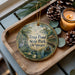 A decorative ornament with a scenic image of a pond and surrounding nature, featuring a bird and a boat, is displayed on a wooden tray alongside pine cones and a lit candle.