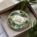 A decorative ornament in a box, featuring a scenic landscape with a waterfall and pine trees.