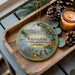A wooden tray with a decorative ornament featuring a scenic lake landscape, pine cones, and a lit candle.