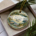 A decorative ornament in a box, featuring a scenic landscape with a lake, trees, and a house.