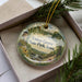 A decorative ornament in a box, featuring a scenic landscape with a castle, trees, and a waterfall.