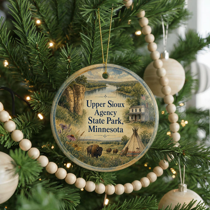 A round ornament hanging on a Christmas tree, featuring a scenic landscape with a river, mountains, and a teepee.