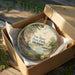 A decorative ornament in a box, featuring a scenic landscape with a lake, trees, and mountains.