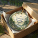 A decorative ornament in a wooden box, featuring a scenic landscape with pine trees and a winding path, with the text "Vaughan Woods State Park, Maine" printed on it.
