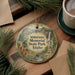 A decorative ornament with a scenic landscape of Veterans Memorial State Park in Idaho, featuring a waterfall and a monument, is displayed on a wooden surface alongside a cup of coffee and some pine branches.
