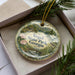 A decorative ornament in a box, featuring a scenic landscape with mountains, a river, and various flora and fauna.