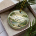 A decorative ornament with a scenic landscape of a river, trees, and a boat, displayed in a box with pine branches.