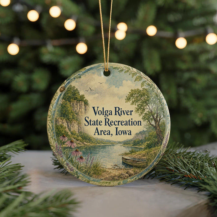 A round ornament with a scenic landscape depicting a river, trees, and a boat, hanging in front of a blurred Christmas tree with lights.