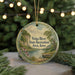 A round ornament with a scenic landscape depicting a river, trees, and a boat, hanging in front of a blurred Christmas tree with lights.