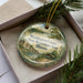 A decorative ornament in a box, featuring a scenic landscape with a river, trees, and a building.
