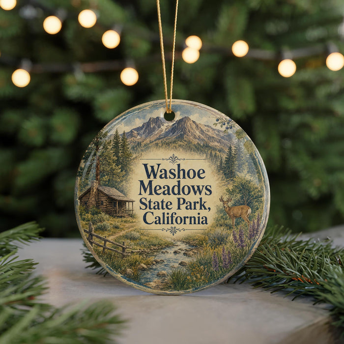 A round ornament with a scenic landscape featuring a cabin, mountains, and a deer, hanging in front of a blurred Christmas tree with lights.