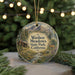 A round ornament with a scenic landscape featuring a cabin, mountains, and a deer, hanging in front of a blurred Christmas tree with lights.
