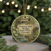 A round ornament with a painting of a bridge and a bird, surrounded by greenery, hangs in front of a blurred background with twinkling lights.