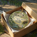 An oval-shaped ornament with a painting of a bridge and a bird, placed in a cardboard box with a gold string.