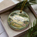 A decorative ornament in a box, featuring a scenic landscape with a bird, flowers, and a body of water.