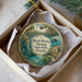 A round ornament with a mermaid and a house, surrounded by greenery, is displayed in a box.