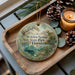A wooden tray with a decorative ornament featuring a scenic image of Wekiwa Springs State Park, Florida, along with pine cones and a lit candle.