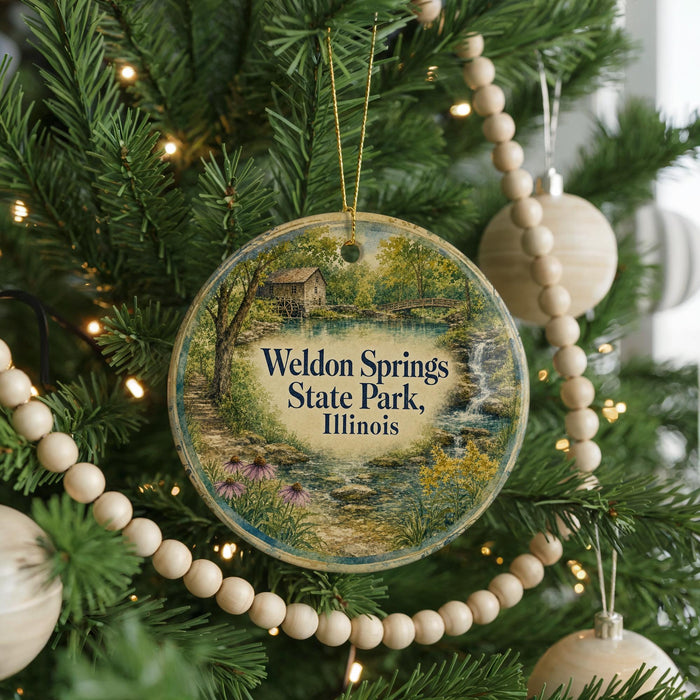 A round ornament hanging on a Christmas tree, featuring a scenic landscape with a house and a stream.