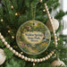 A round ornament hanging on a Christmas tree, featuring a scenic landscape with a house and a stream.