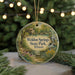 A round ornament with a painting of a scenic landscape, featuring a cabin and a stream, hangs in front of a blurred background with twinkling lights.