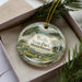 A decorative ornament in a box, featuring a scenic landscape of Wells State Park in Massachusetts.