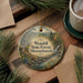 A round ornament with a scenic forest landscape, featuring pine trees, a waterfall, and mountains, is displayed on a wooden surface alongside a cup of coffee and some papers.