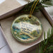 A round ornament with a scenic image of a pond and wildlife, including a beaver and a crane, is displayed in a box with pine branches.