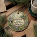 A decorative ornament with a scenic illustration of White Clay Creek State Park in Delaware, featuring a bird, flowers, and a house, is displayed on a wooden surface alongside a cup of coffee and some pine branches.