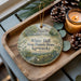 A decorative ornament featuring a house and trees, placed on a wooden tray next to a lit candle and pine cones.