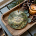 A decorative ornament featuring a scenic landscape of White Pines Forest State Park, Illinois, with pine cones and a lit candle on a wooden tray.