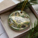 A decorative ornament in a box, featuring a scenic landscape with a waterfall and greenery, with the text "Wildcat Den State Park, Iowa" prominently displayed.