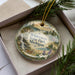A decorative ornament with a landscape scene featuring a deer, pine trees, and a waterfall, placed inside a box with pine branches.
