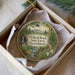 A round ornament with a scenic forest landscape, featuring a winding path, waterfall, and pine trees, is displayed in a box.