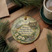 A decorative ornament featuring a house and flowers, with the text "William Whitley House State Historic Site, Kentucky" printed on it. The ornament is hanging on a wooden surface, surrounded by pine branches and a cup of coffee.