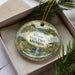 A decorative ornament in a box, featuring a scenic landscape with a waterfall, pine trees, and a deer.