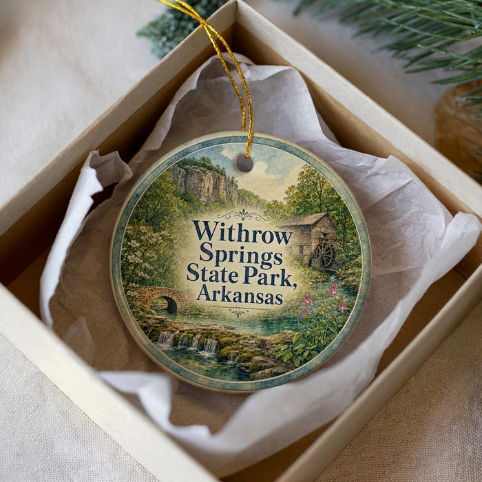 A decorative ornament with a scenic landscape of springs, state park, and a small building, placed in a box with a white tissue paper lining.